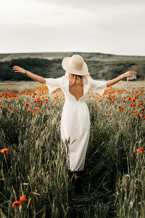 A lady strolling through a field full of flowers while wearing a white summer dress and hat
